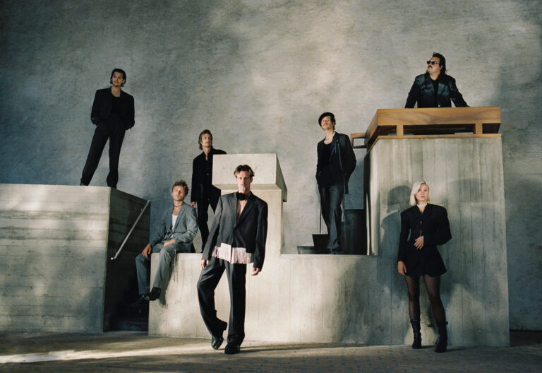 Bandfoto van Son Mieux, de 7 leden staan in een studio met een grijze achtergrond en blokken, leden zitten op de blokken en staat er tegenaan. Ze kijken naar de camera en om zich heen.