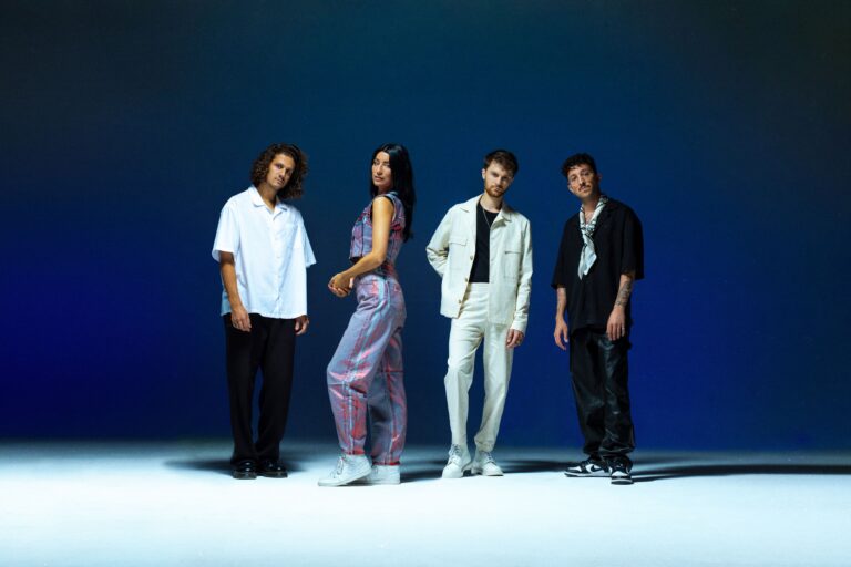 Een foto van de band rondé, de 4 leden staan voor een donkerblauwe muur en op een lichtblauwe vloer en kijken naar de camera.