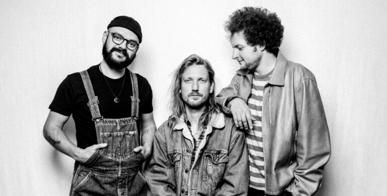 De drie mannen van Subterranean Street Society staan voor een witte muur, twee kijken in de camera en 1 kijkt naar links.