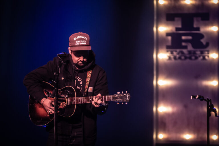 Een muzikant met een gitaar staat op het podium, verlicht door blauwe en warme lichten.