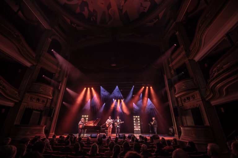 Een band staat op een podium in een theater, omringd door kleurrijke lichten met een publiek in de verte.
