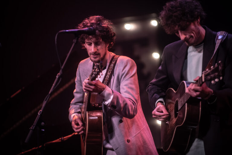 Twee muzikanten spelen gitaar op het podium met een sfeervolle achtergrondverlichting.