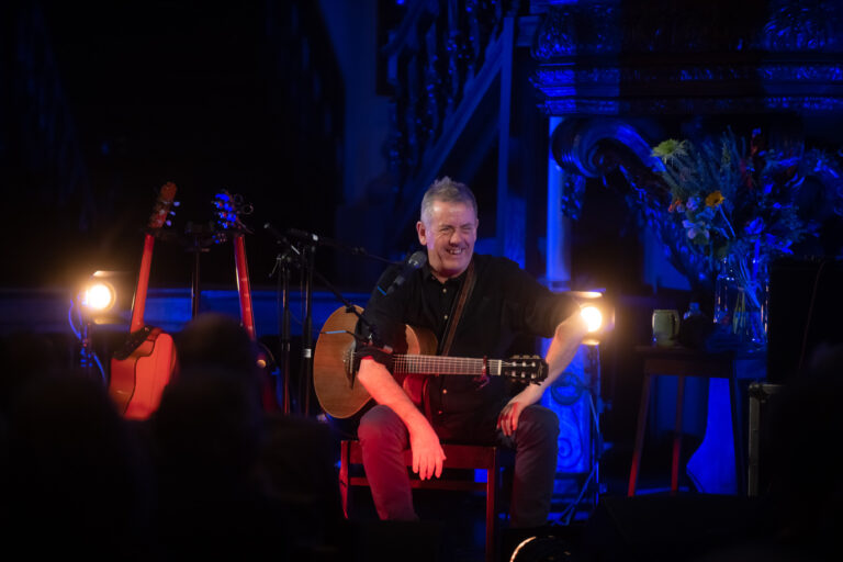 Een muzikant zit op een stoel met een gitaar in een sfeervol verlichte ruimte.