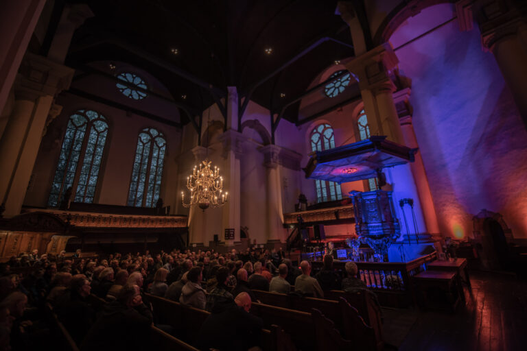 Een drukbezochte kerkzaal met veel mensen, sfeervolle verlichting en grote ramen.