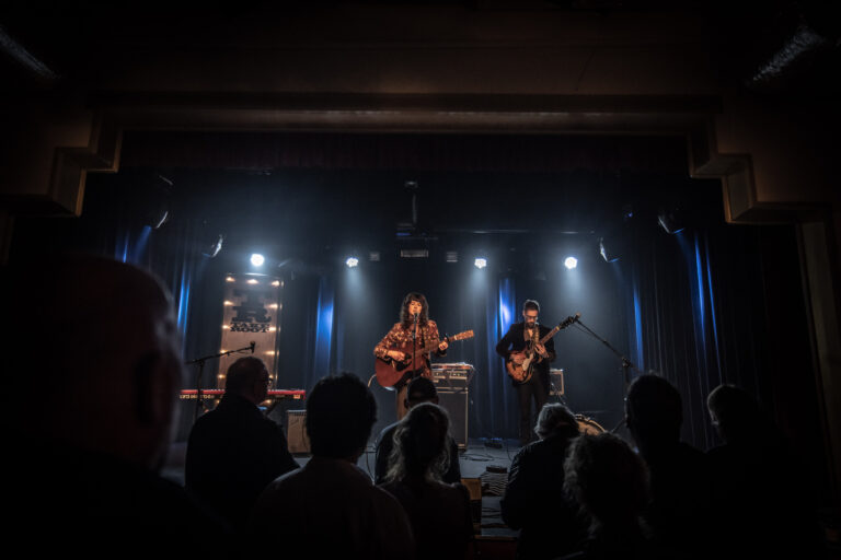 Een live muziekoptreden met twee musicians op het podium, verlicht door schijnwerpers, terwijl het publiek toekijkt.