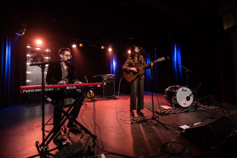 Een muzikale uitvoering met een zangeres die gitaar speelt en een muzikant achter een keyboard, verlicht door sfeervolle lichten.