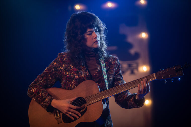 Een muzikant met krullend haar speelt gitaar op een sfeervol verlichte podium.