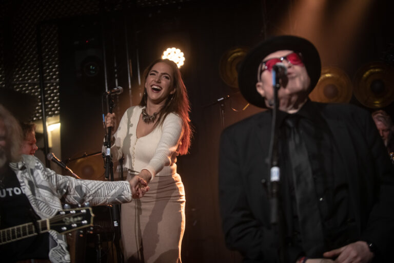 Een muzikale optredengroep met een zanger en een zangeres op het podium, omringd door sfeerverlichting.