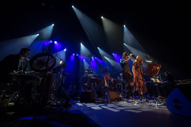 Een muzikale uitvoering met twee zangers op het podium, omringd door muzikanten en verlicht door blauwe en paarse lichten.