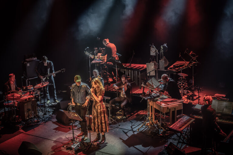Een live muziekopvoering met meerdere muzikanten op het podium, waaronder een zangeres en verschillende instrumenten.