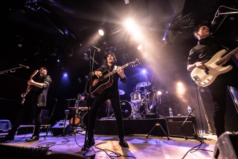 Een band speelt live op het podium met een gitarist in het midden, omringd door bandleden en sfeervolle lichten.