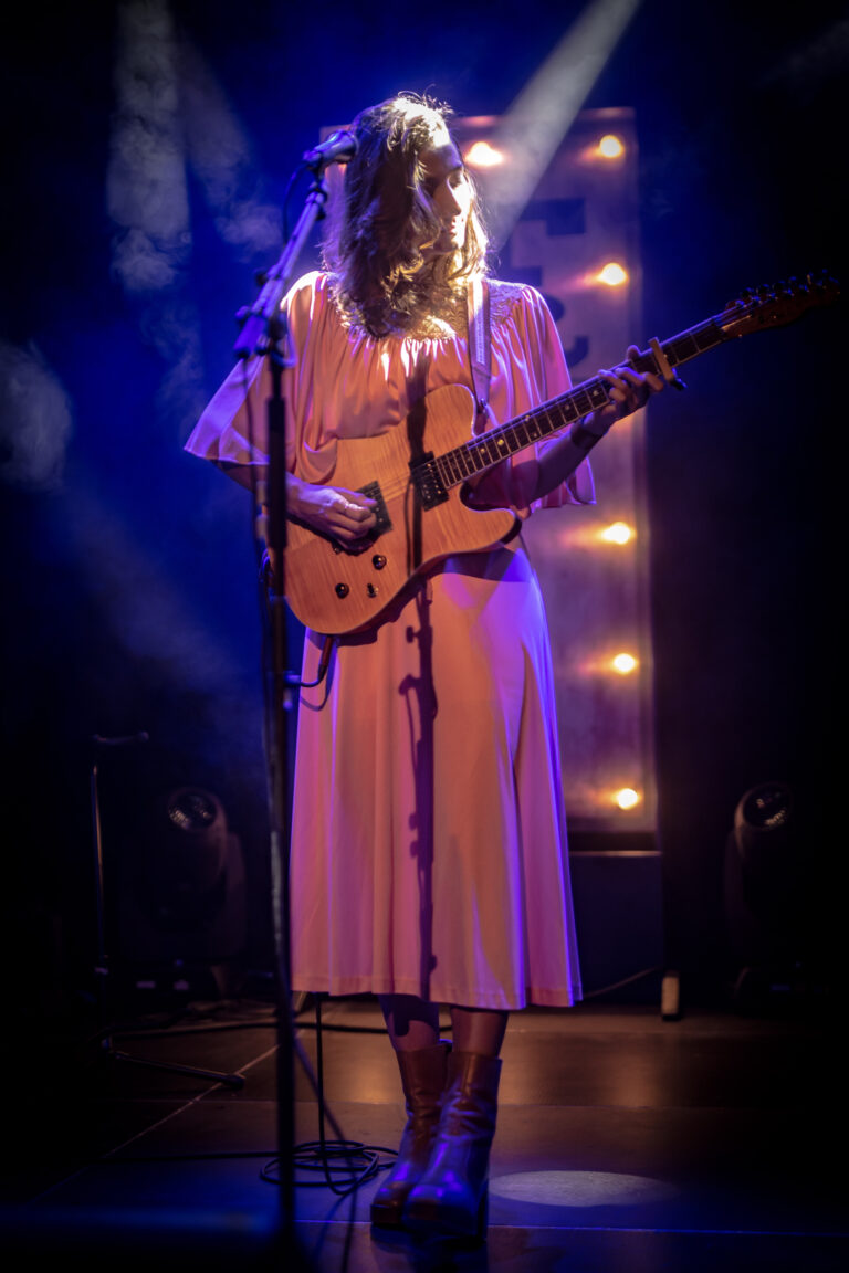 Een muzikante speelt gitaar op het podium, omgeven door sfeervolle verlichting.