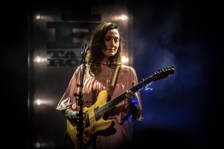 Een muzikante speelt op een elektrische gitaar tijdens een optreden, omgeven door sfeervolle verlichting.