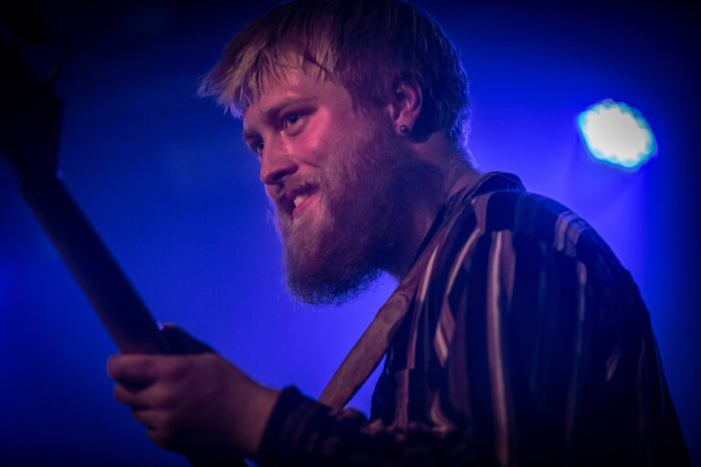 Een muzikant met een baard speelt een gitaar op een podium, omgeven door blauwe verlichting.