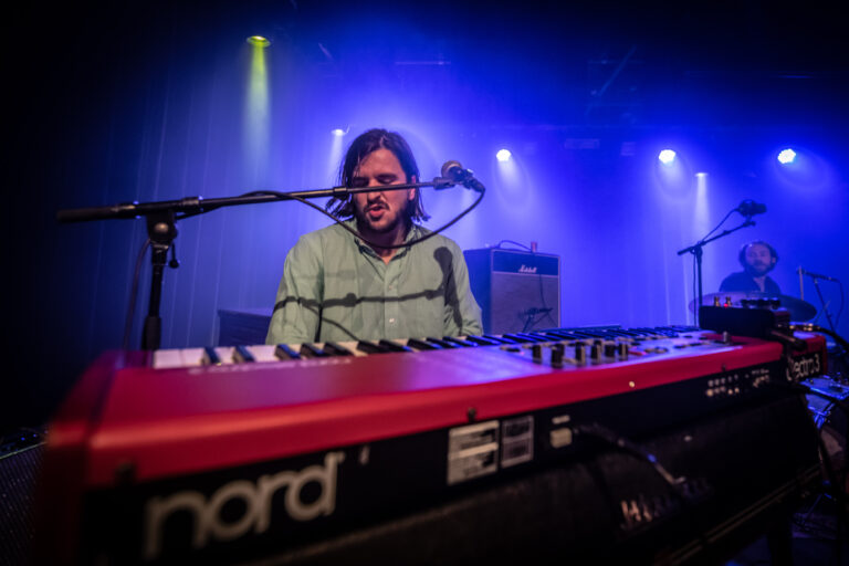 Een muzikant speelt op een rode digitale piano tijdens een live optreden met blauwe en gele lichteffecten op de achtergrond.