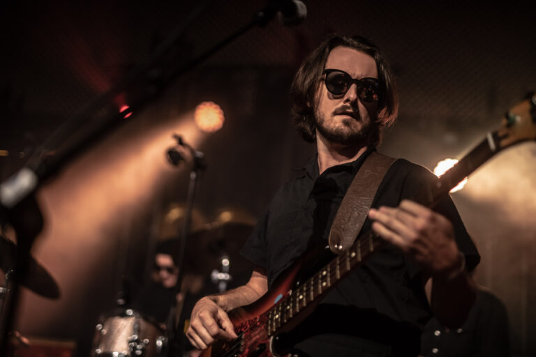 Een muzikant met een gitaar op het podium, gekleed in zwart en met een zonnebril, terwijl hij optreedt in een sfeervolle setting.