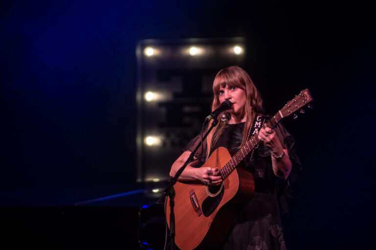 Een muzikante zingt en speelt gitaar op een podium, omringd door dim licht en een retro achtergrond.