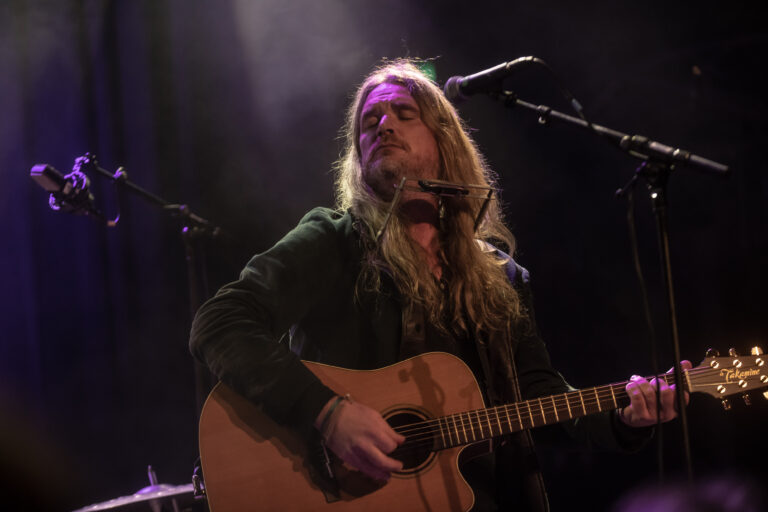 Een muzikant met lang haar speelt gitaar op een podium met zachte verlichting.