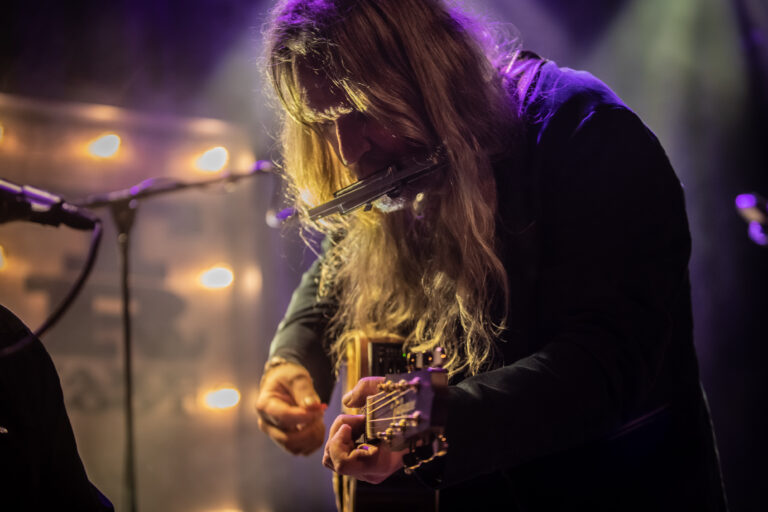 Een muzikant met lang haar speelt gitaar en harmonica op een sfeervol verlichte podium.