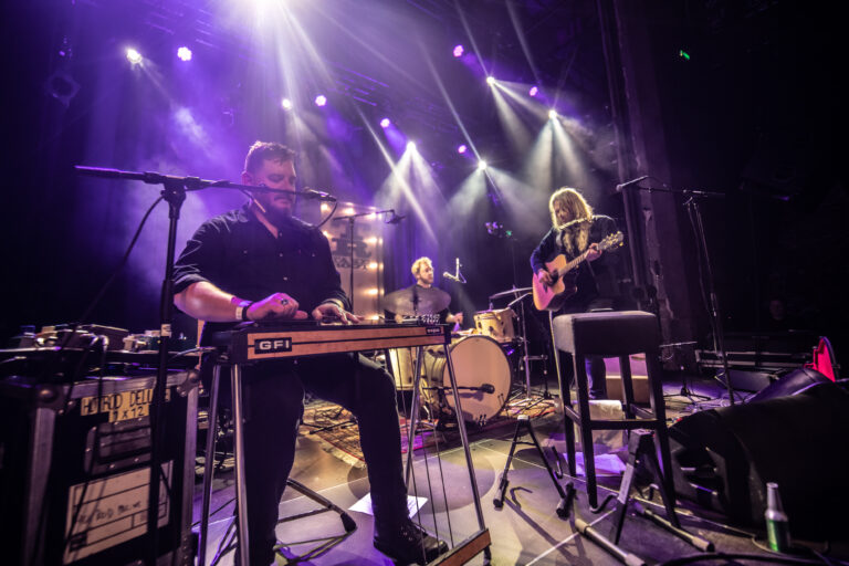 Drie muzikanten spelen op een podium met kleurrijke verlichting en een uitgelichte achtergrond.