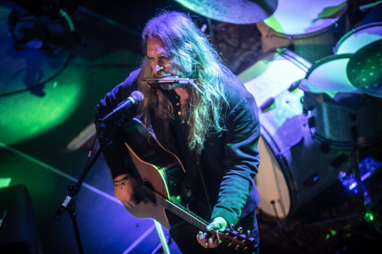 Een muzikant met lang haar speelt gitaar en zingt in een sfeervolle, verlichte omgeving.