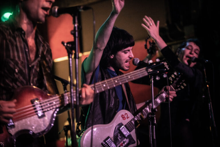 Een muzikant met een gitaar zingt in een levendige concertsetting terwijl andere bandleden op de achtergrond hun instrumenten bespelen.