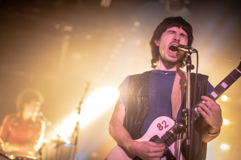 Een muzikant zingt vol energie in een concertomgeving met opvallende lichtstralen.