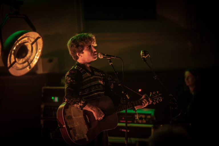 Een muzikant met een gitaar zingt op een podium, omringd door sfeerverlichting.
