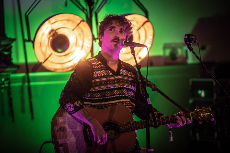 Een muzikant met een gitaar performt op het podium, omringd door sfeervolle lichten.