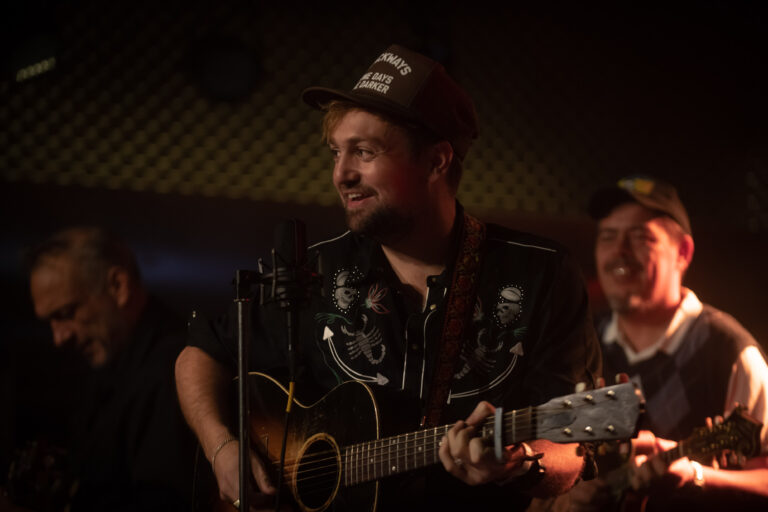 Een gitarist speelt en zingt met een glimlach in een muzikale setting, omringd door andere muzikanten.