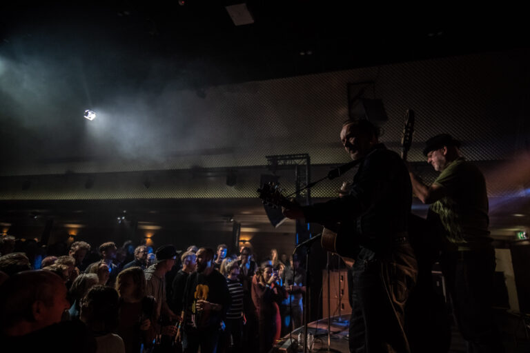 Een band voert op een podium live muziek uit voor een enthousiast publiek in een sfeervolle setting met lichteffecten.