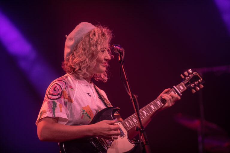 Een muzikant met krullend haar en een hoed speelt gitaar op een kleurrijk verlichte podium.