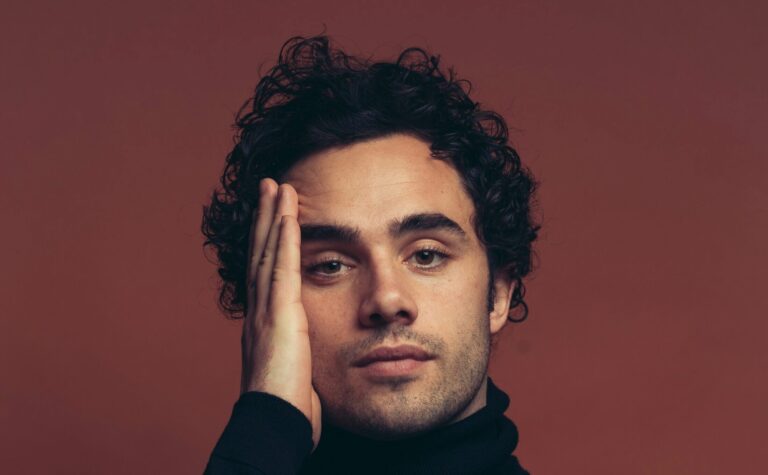 Portretfoto van zanger Toby Sebastian, hij zit voor een donkerrode muur en heeft een zwarte coltrui aan, hij houdt zijn hand tegen zijn gezicht.
