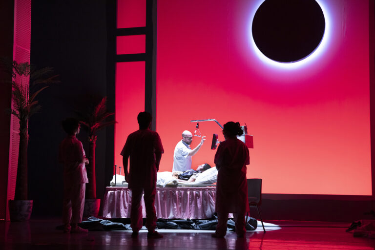 Een man staat achter een tafel in een theater, omringd door silhouetten van bezoekers, met een heldere rode achtergrond en een ringvormig object dat lijkt op een zon of maan.