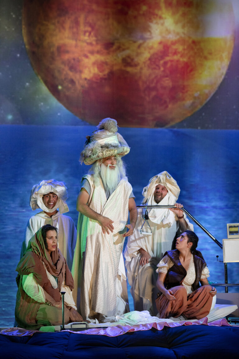 Een theatrale voorstelling met vijf acteurs in kostuums, tegen een achtergrond van een grote planeet en een zee.