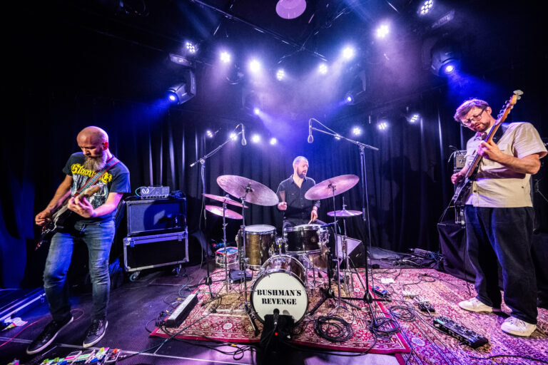 Een band speelt live op het podium met een drumstel en elektrische gitaren, omgeven door kleurrijke lichten.