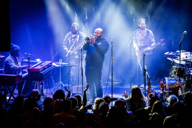 Een live band speelt op het podium, omgeven door kleurrijke lichtstralen en een enthousiaste menigte.