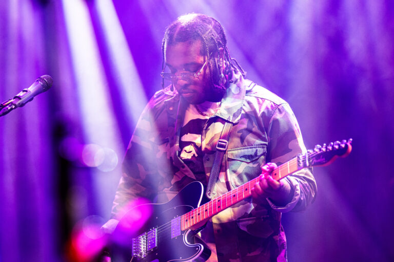 Een muzikant speelt gitaar op een podium, omgeven door kleurrijke lichtstralen.