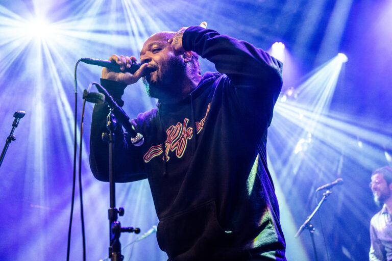 Een muzikant zingt met een microfoon op een podium, omringd door kleurrijke lichtstralen.