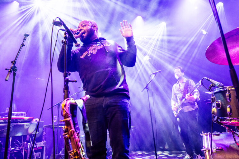 Een zanger performt op het podium met een saxofonist, omringd door kleurrijke lichtstralen.