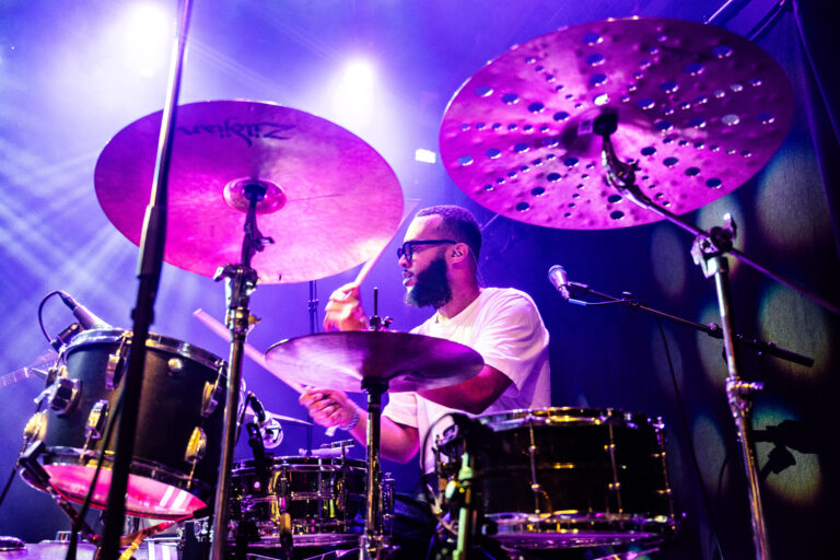 Een drummer speelt op een podium onder kleurrijke verlichting met grote cymbalen en een drumstel.