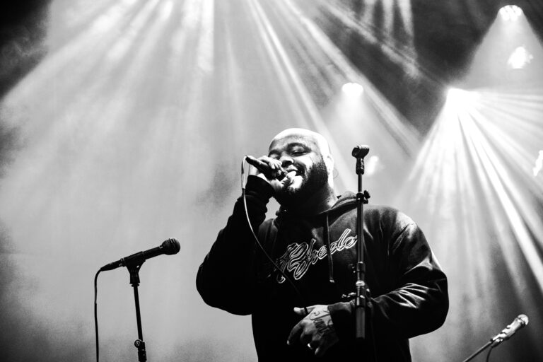 Een zanger staat op het podium en zingt in een verlichte, rookachtige omgeving.