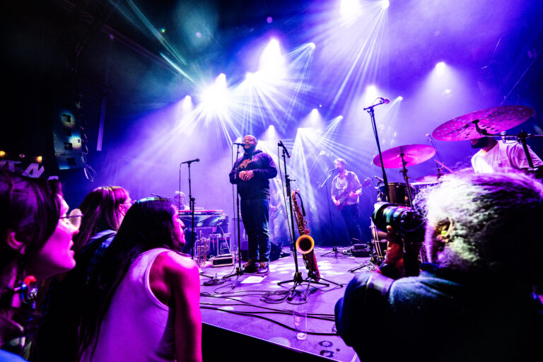 Een muzikant staat op het podium omgeven door kleurrijke lichten, terwijl het publiek aandachtig luistert.