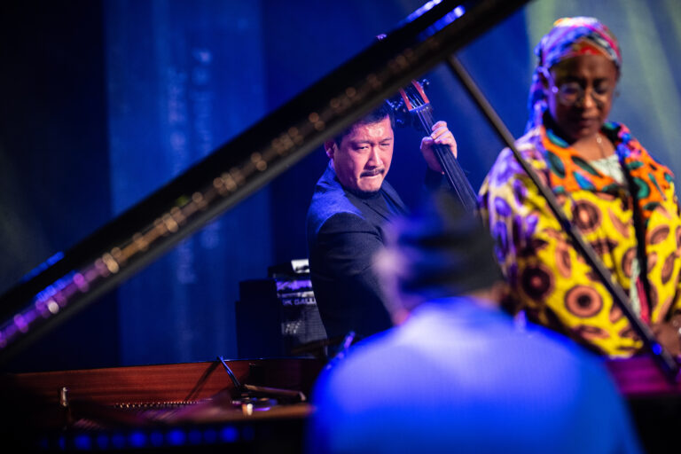 Een muzikant speelt contrabas op een podium, met een piano en andere muzikanten op de achtergrond.