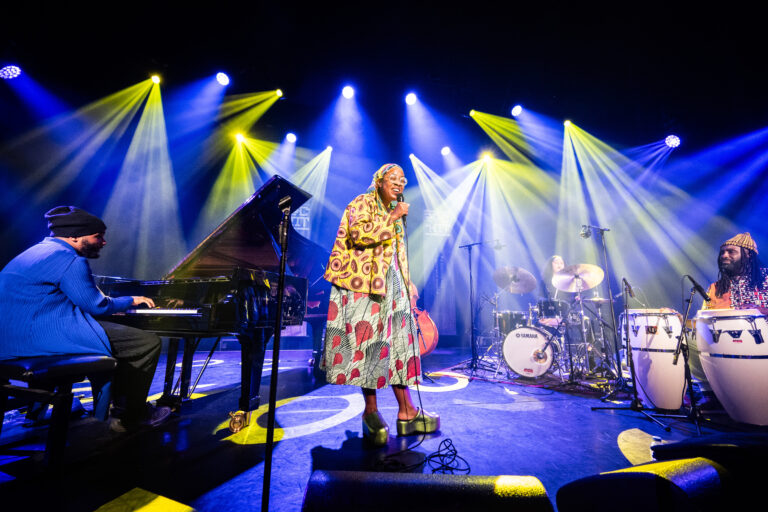 Een zangeres met een kleurrijke jurk staat op het podium terwijl ze een live optreden geeft, omringd door muzikanten en sfeerverlichting.
