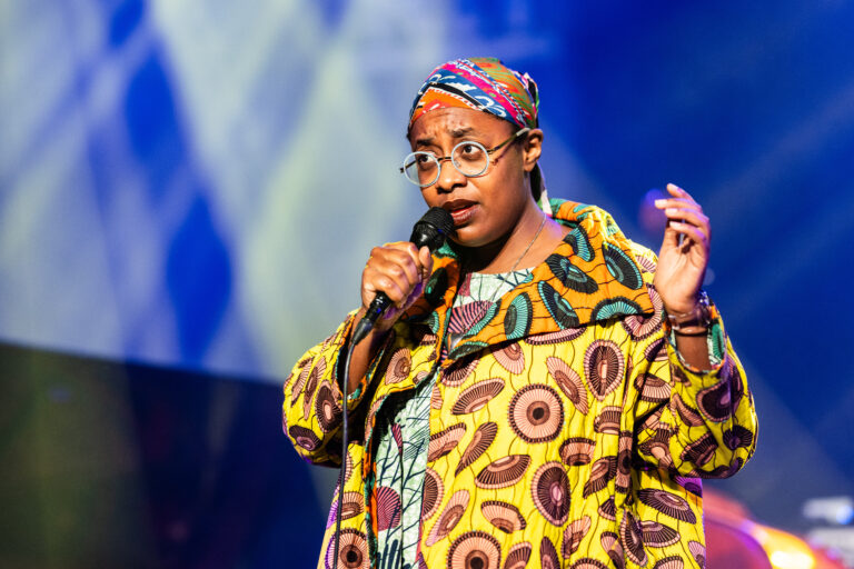 Een persoon in een kleurrijke, traditional outfit houdt een microfoon vast terwijl ze op het podium staat.