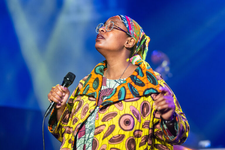Een vrouw in kleurrijke kleding staat op een podium met een microfoon in haar hand en kijkt omhoog.