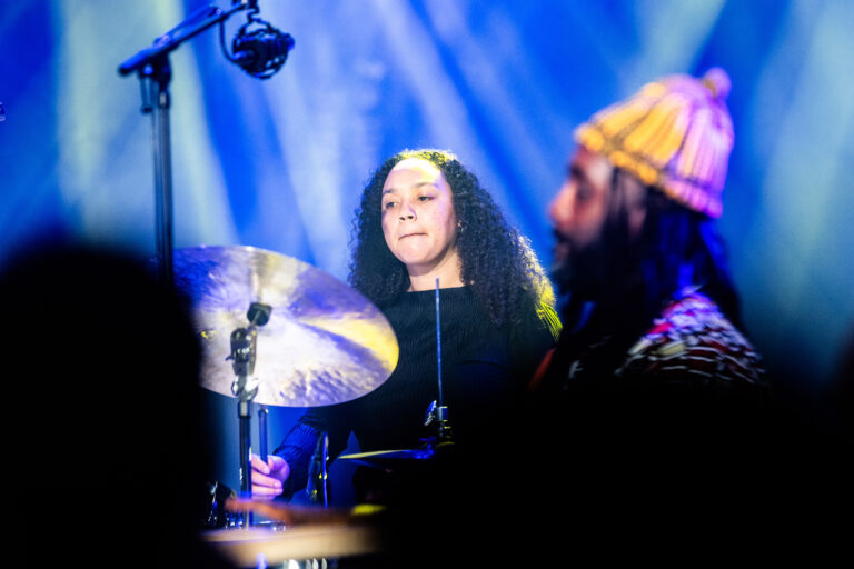 Een vrouwelijke drummer speelt op een podium met kleurrijke verlichting.