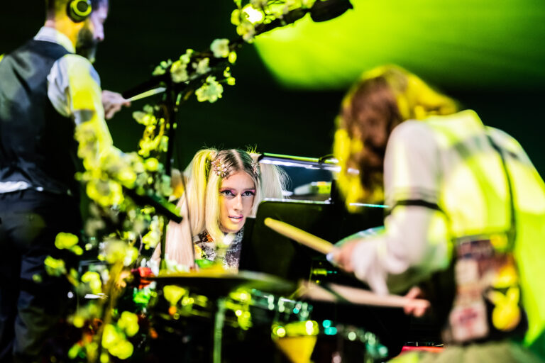 Een drummer wordt gefotografeerd terwijl ze achter haar instrument zit, omringd door bloemen en sfeerverlichting.