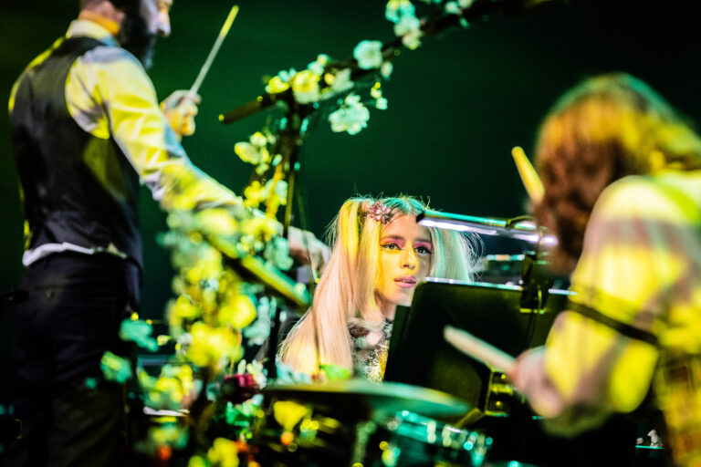 Een muzikante speelt op een drumstel omringd door bloemen tijdens een optreden.
